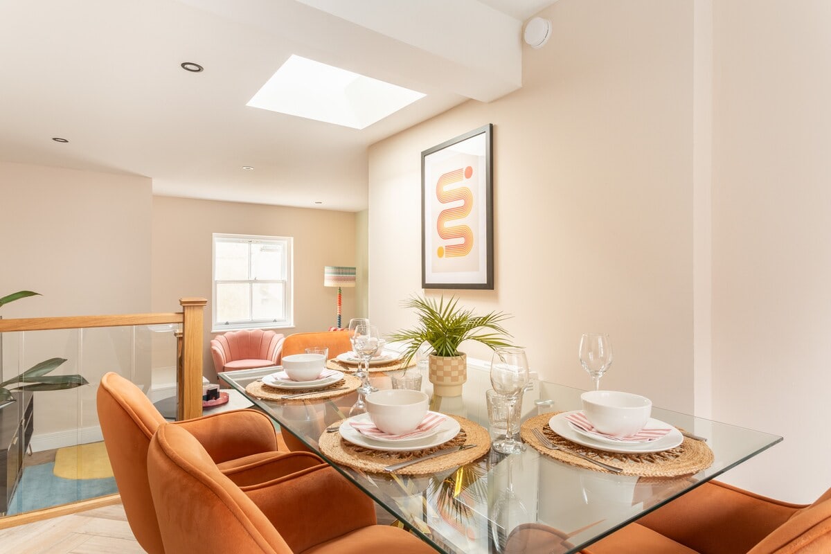 Glass dining table with orange chairs beneath a skylight beside a glass stair balustrade